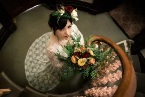 stunning boho wedding bridal in lace vintage bridal gown standing at the bottom of a fairytale castle grand staircase holding a bunch of greenery woodland flower bouquet smile at the camera