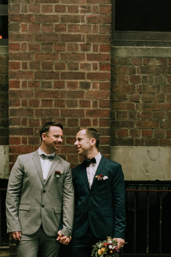 same sex couple happily laughing in front of a brick wall at Hardware Lane wedding photo