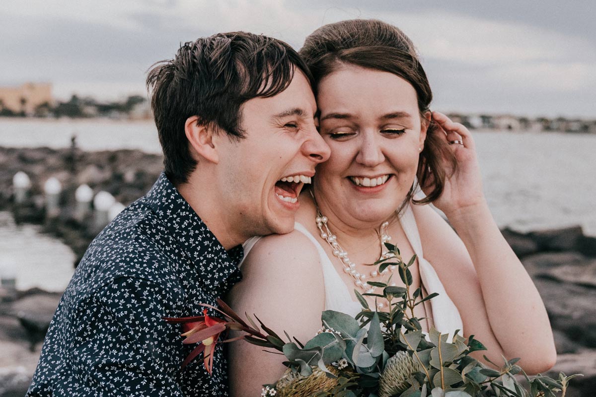 Laughing couple by the St Kilda beach on their wedding day capture by wedding photography company Black Avenue Productions