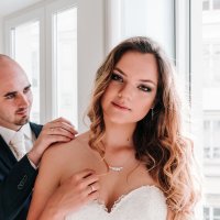 beautiful blonde hair bride Marta look at camera for Europe photo shoot with husband Stefan help her wearing wedding necklace in Vienna apartment