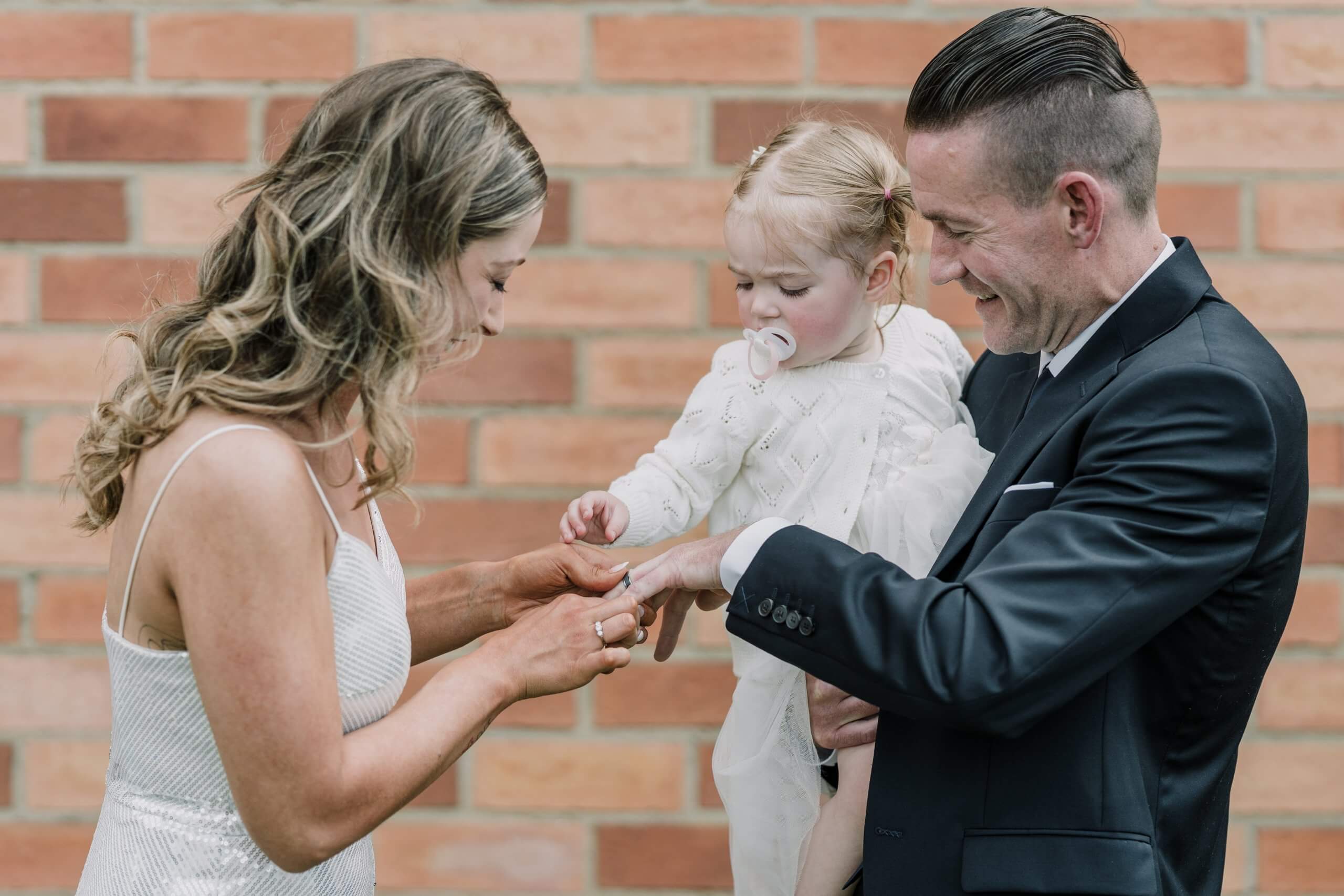 The lovely bride putting the wedding ring to the groom's finger while he's carrying their lovely daughter, captured by Black Avenue Productions