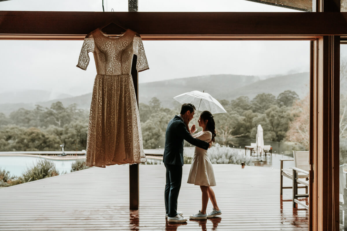 A beautiful shot of the couple standing by the front of the porch while holding an umbrella under the rain Wedding Day Interrupted by bad weather captured by Black Avenue Productions