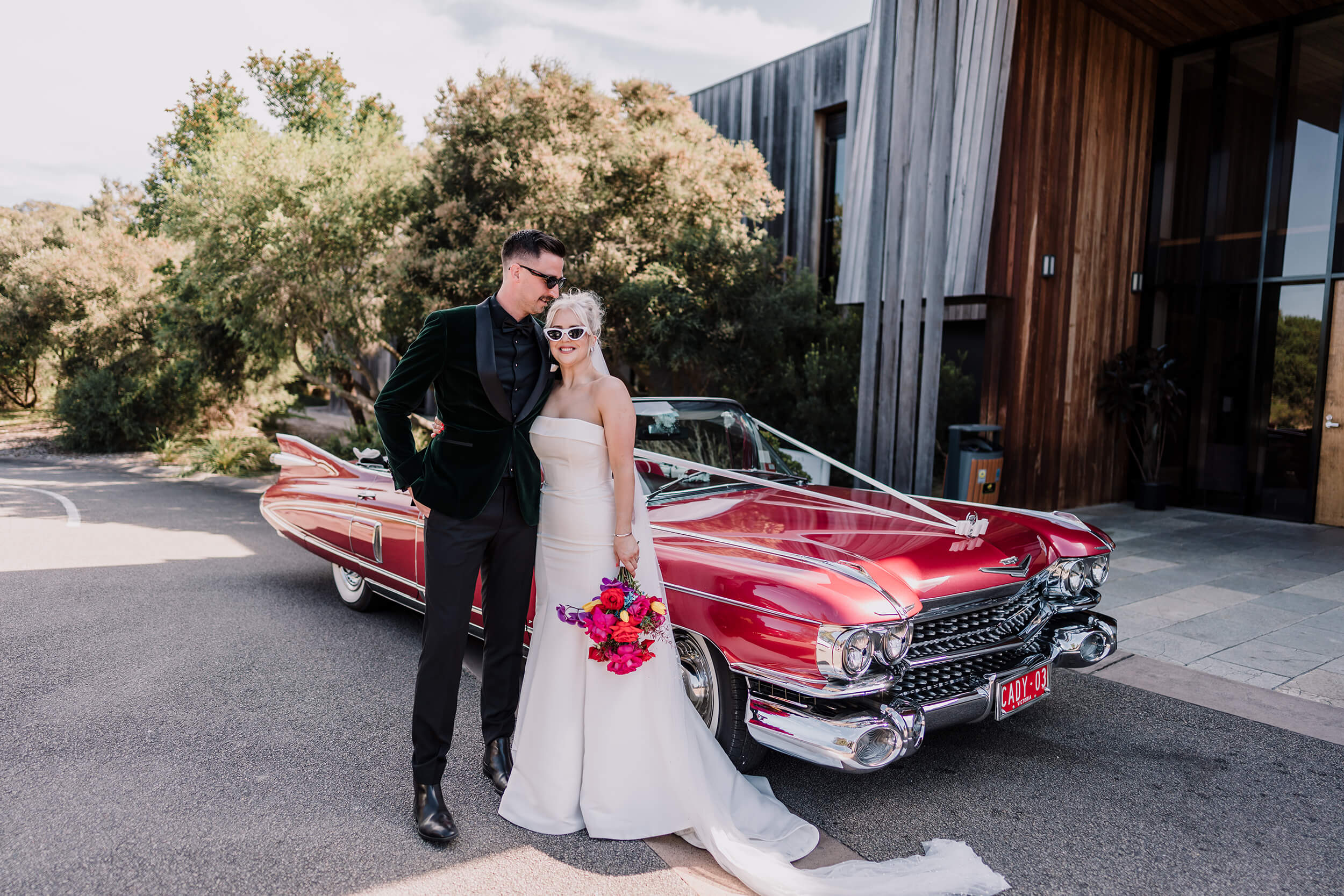 Bride and groom happily posing with shades on beside a red convertible cadillac as their wedding car. Captured by Black Avenue Productions
