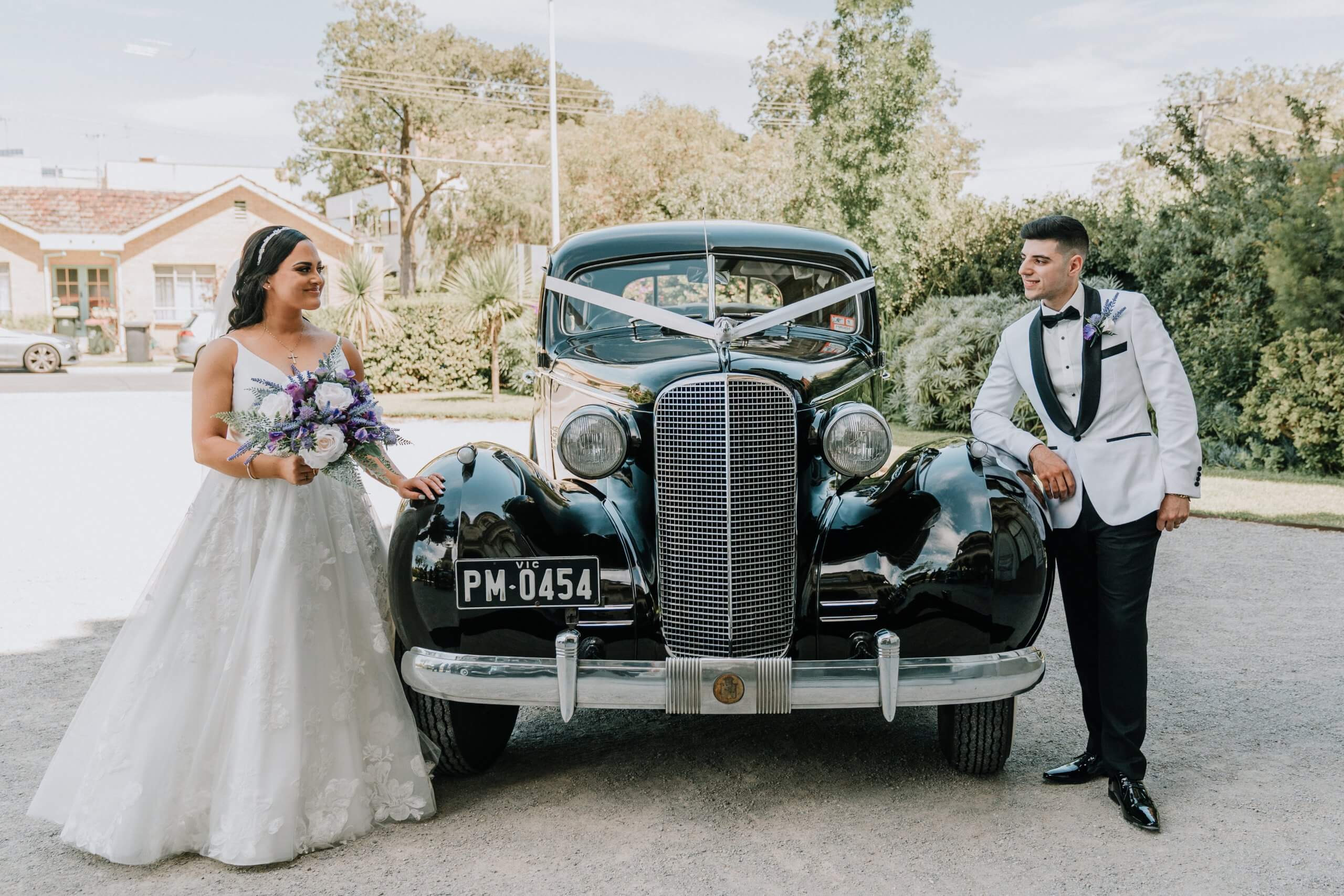 Bride and groom looking in love staring at each other while leaning on the wedding car. Captured by Black Avenue Productions
