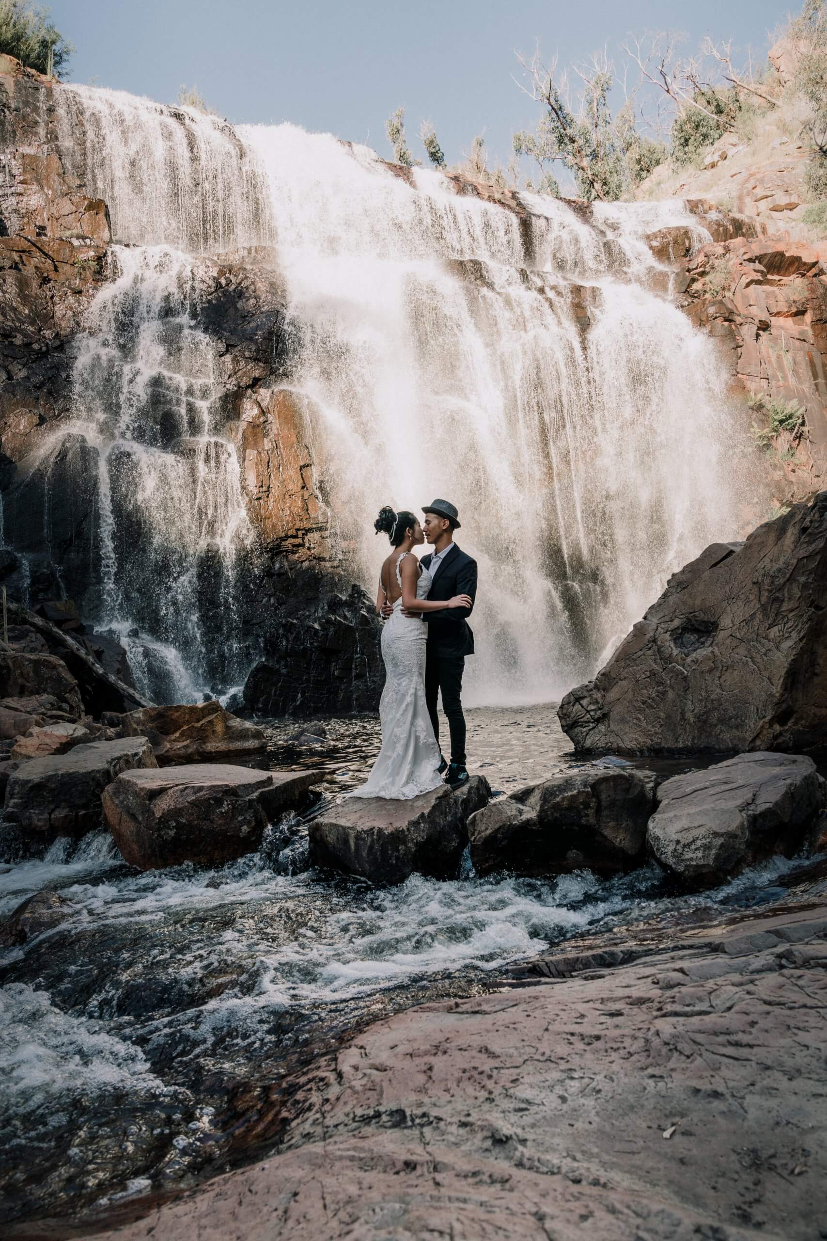 Bride and groom holding each other while standing on a rock in front of a majestic waterfall.