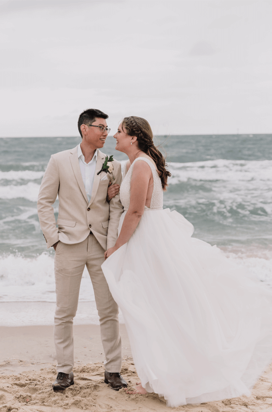 Bride holds on to the grooms arm while they are posing for a photo in the coastline.