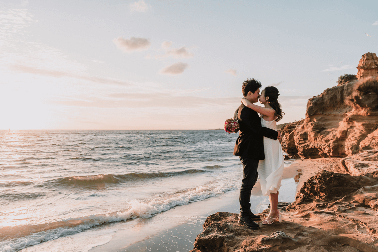 A sunset photo of a bride and groom sharing a hug while standing on the rocky shores of a calm sea.