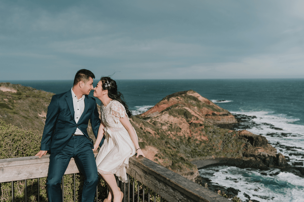 Noses touching, the bride and groom sits on the wooden railings of Cape Schanck with the beautiful view of the sea and hills at the back.