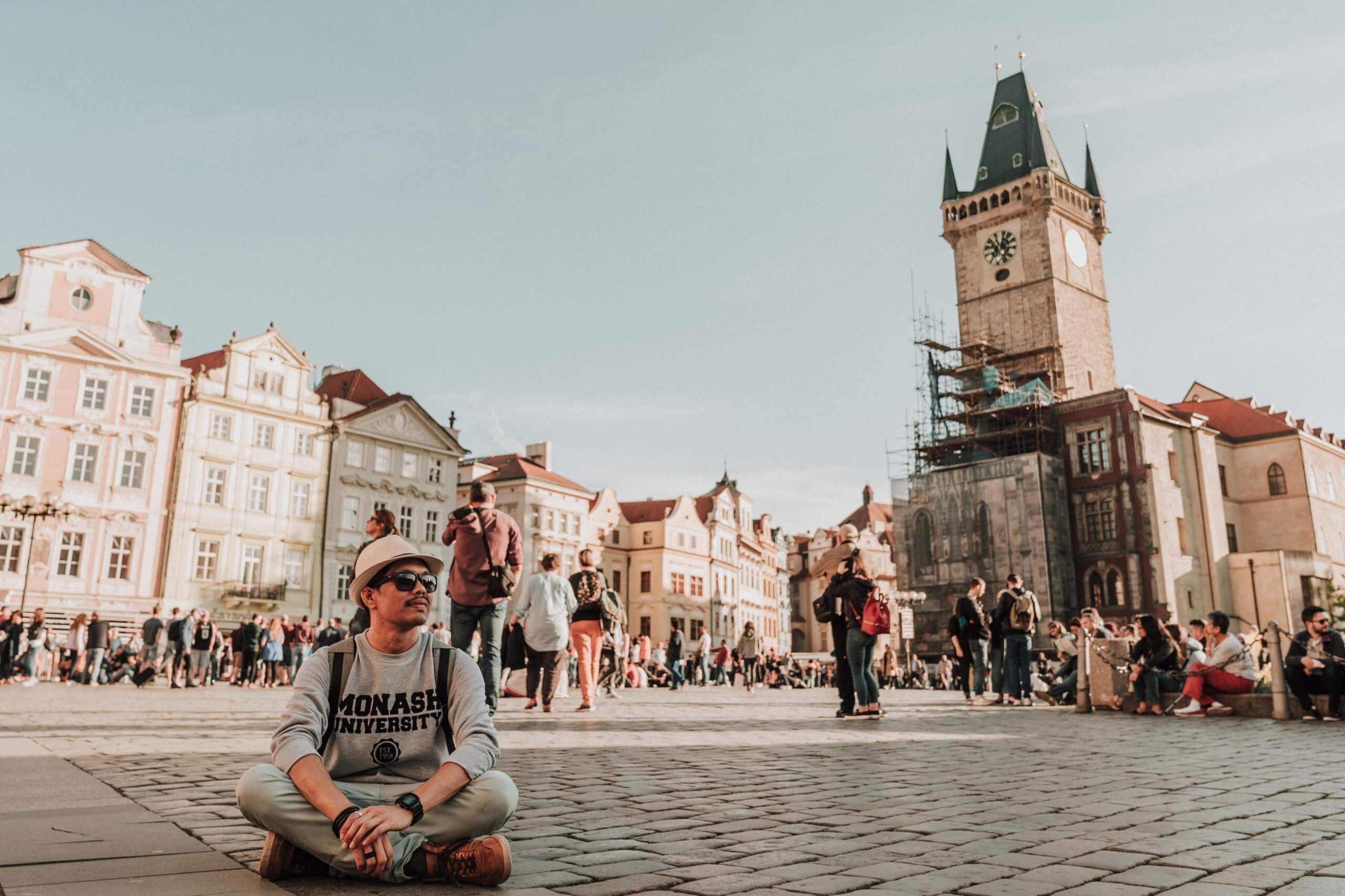 Male photographer sits on the cobblestone floors of Prague with a view of the busy old town square at the back.