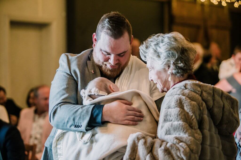 Beautiful photo of a father holding his sleeping baby in his arms while the grandma looks fondly at the child during the ceremony.