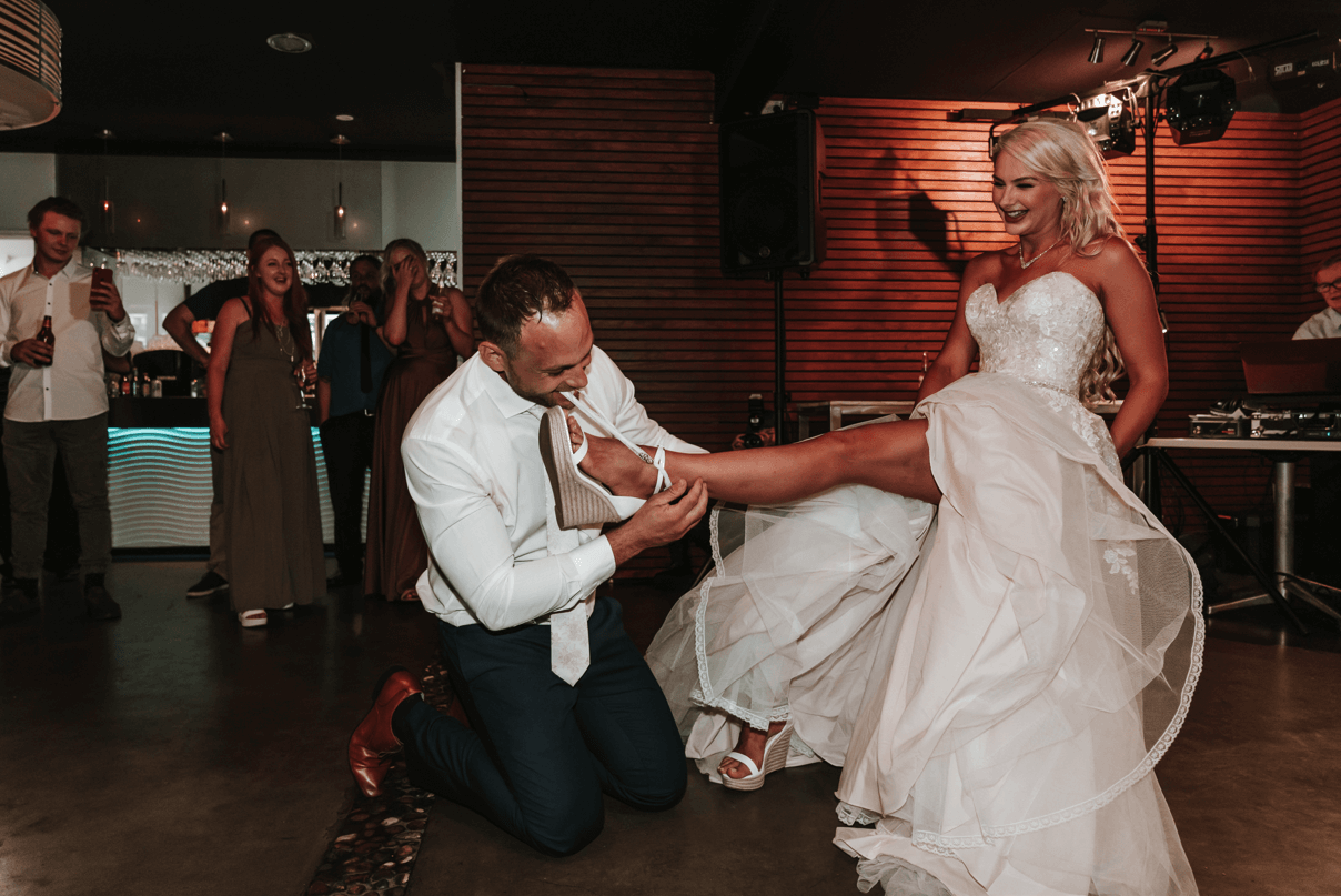 Grooms kneels on the floor as he takes the garter off the bride's leg.