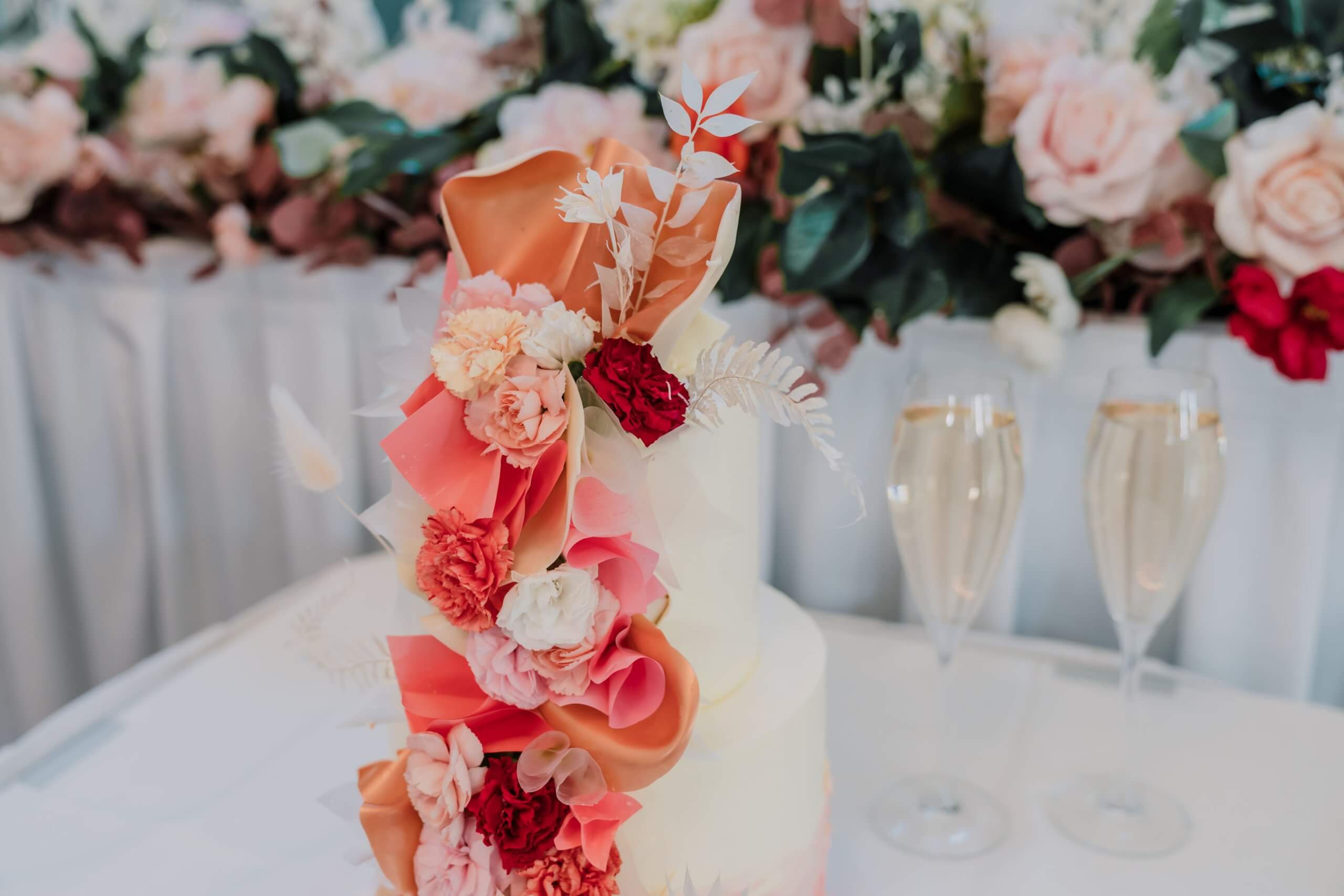 White frosted wedding cake with orange, pink, red, and burgundy flowers.