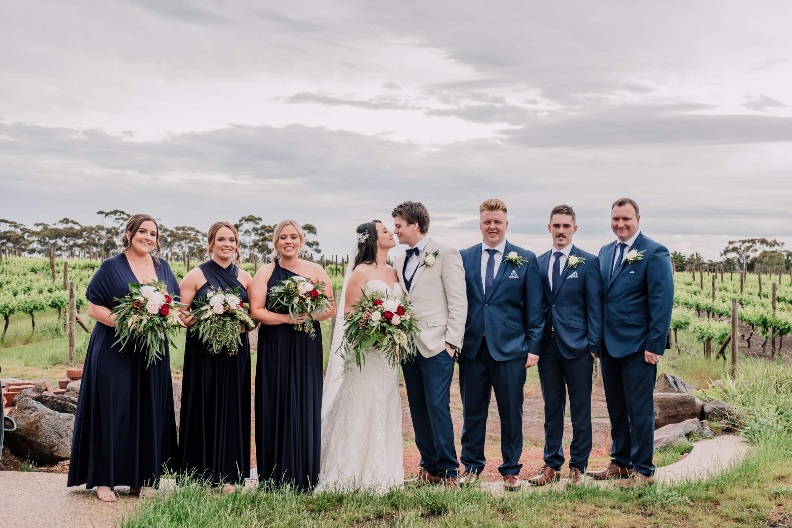 A stunning bridal party photo captured at dusk. The bride and groom, along with their bridesmaids and groomsmen - wearing midnight blue, are surrounded by the soft and warm colors of the setting sun.