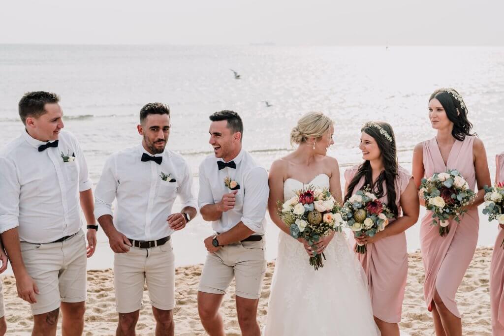A beautiful photo of the bridal party as they stand in front of the shoreline with the setting sun and seagulls flying in the background.