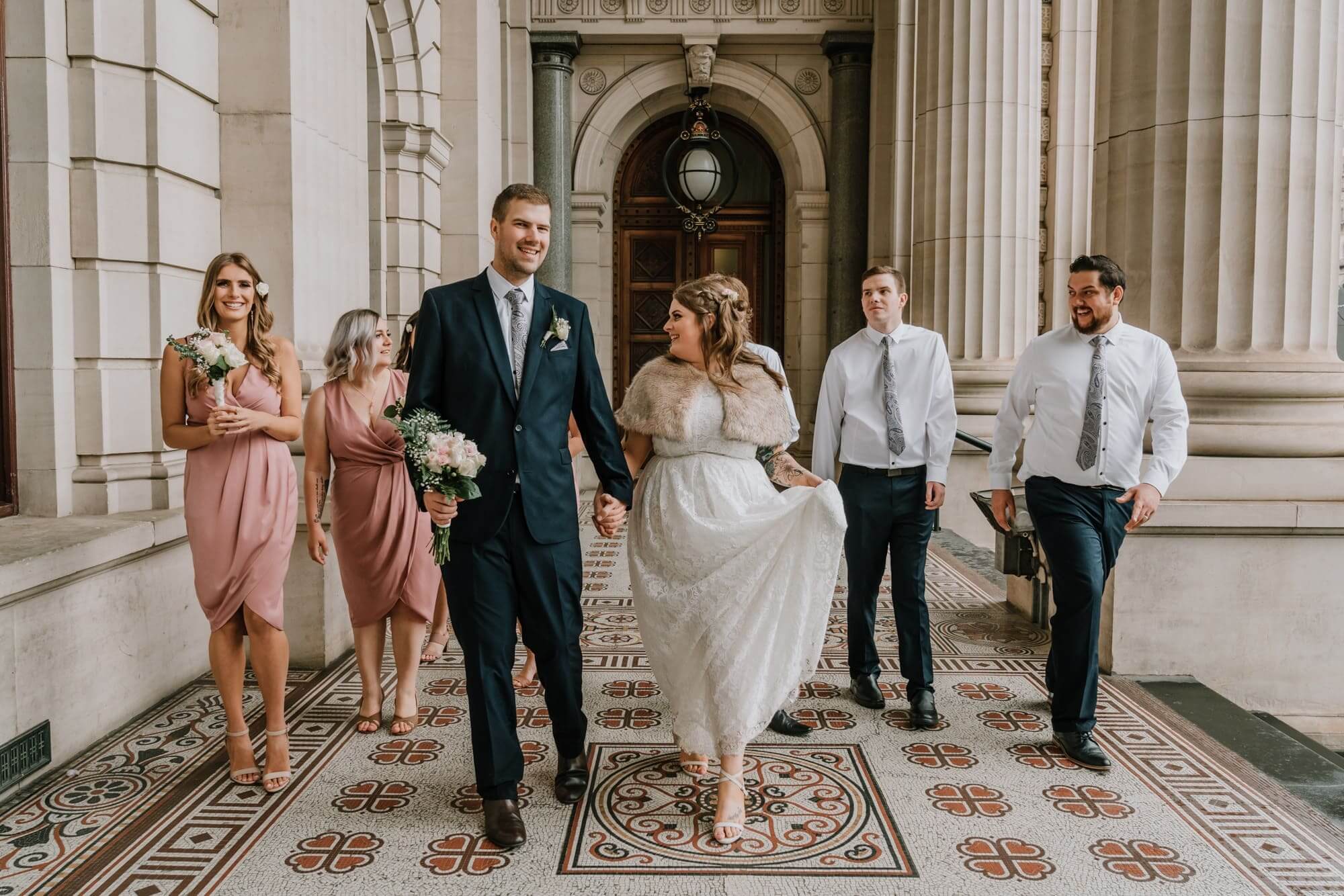 The bride and groom walks along the hallway of a building with large columns - flanked by their bridesmaids and groomsmen in either side.