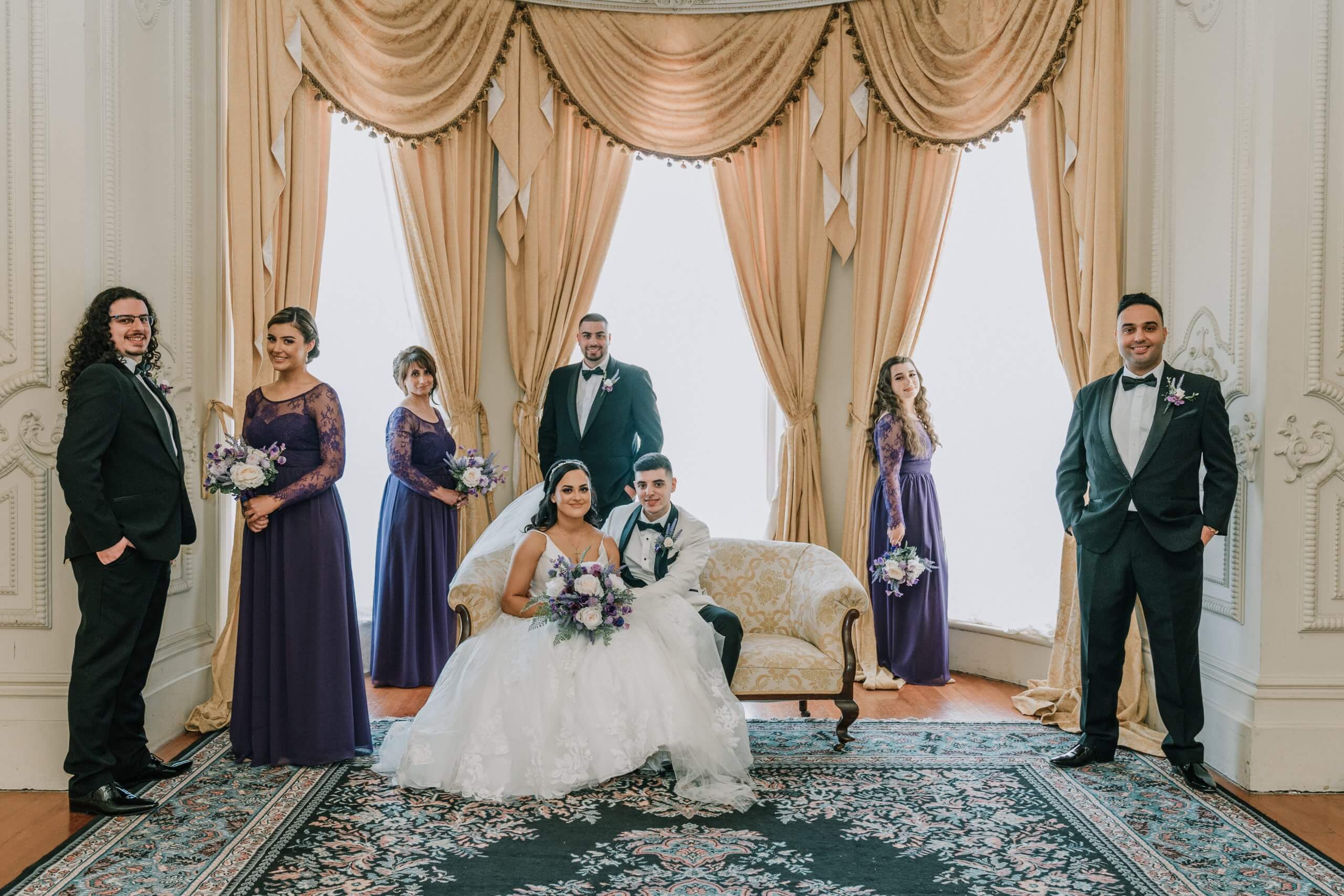A regal photo of the bride and groom with their family. In a carpeted room with high ceilings and windows, couple sits in a love seat while their bridal party, clad in purple and black poses for the photo.