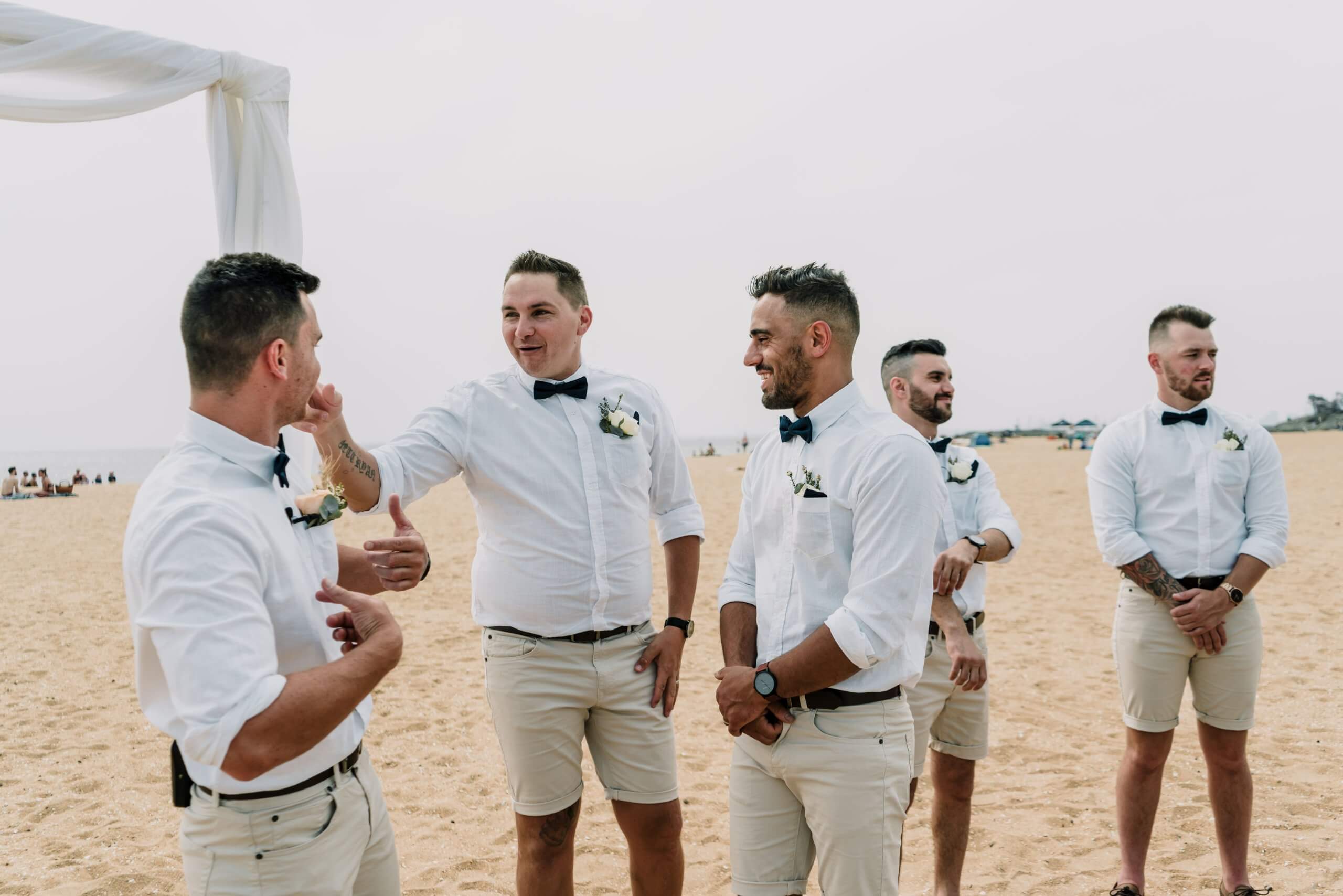 The groom and his groomsmen are being playful in front of the wedding arc as they are waiting for the bride to walk down the sandy aisle of their beachfront wedding.