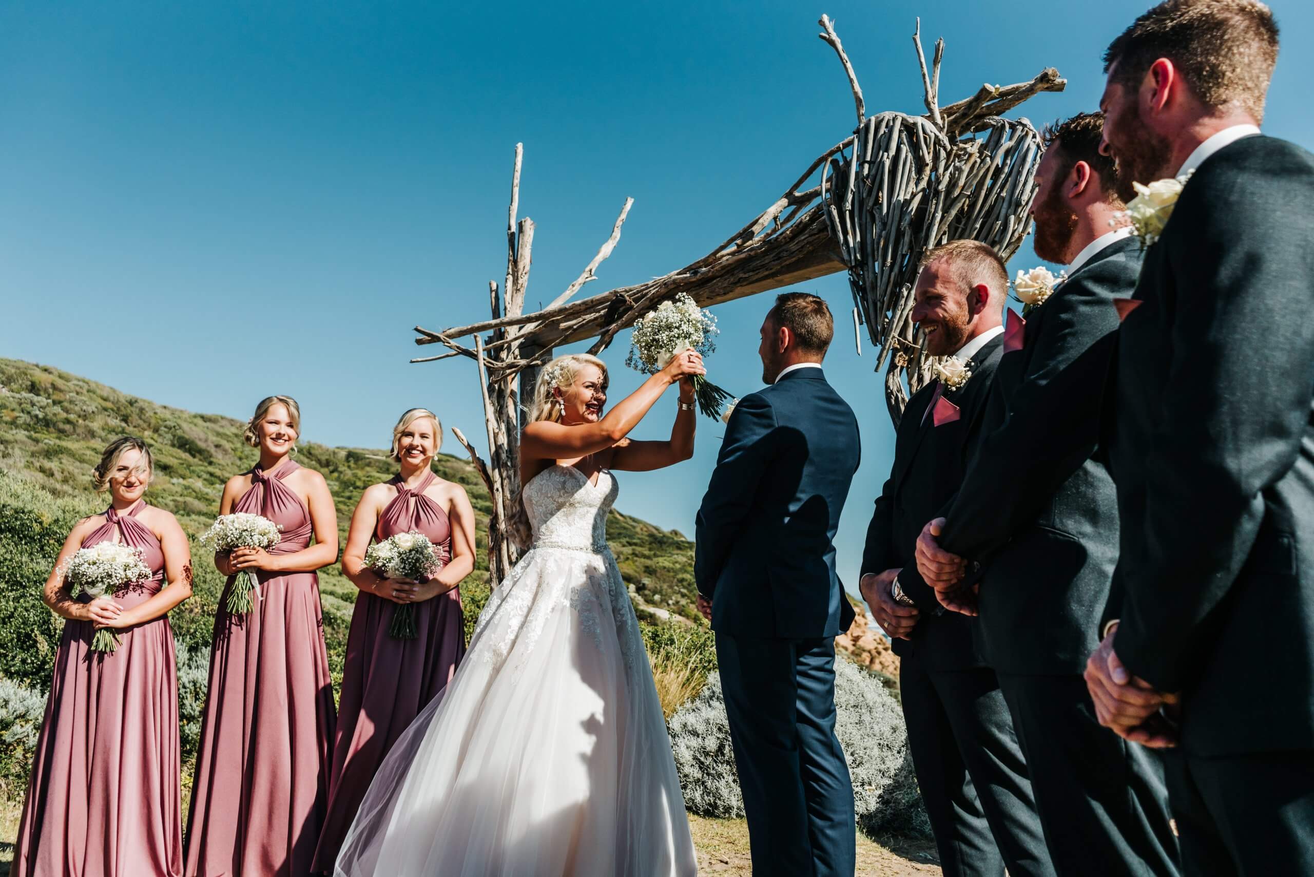The bride playfully shields her face from the sunlight during her beach wedding ceremony where she is standing under the clear blue sky and wedding arch, while being surrounded by her bridesmaids, groomsmen, and groom.