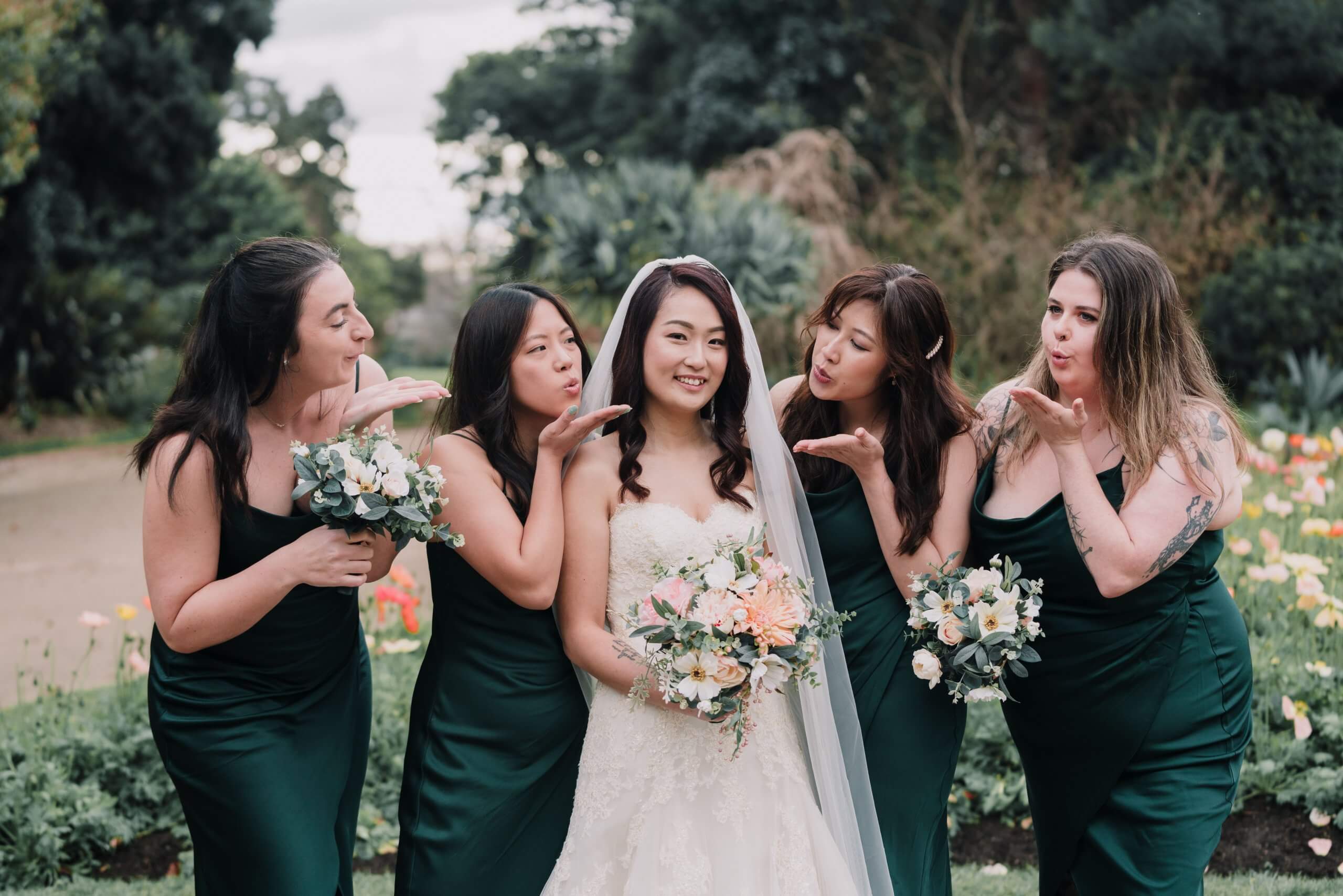 Four bridesmaids, dressed in metallic emerald green dresses, blows kisses on the smiling bride.