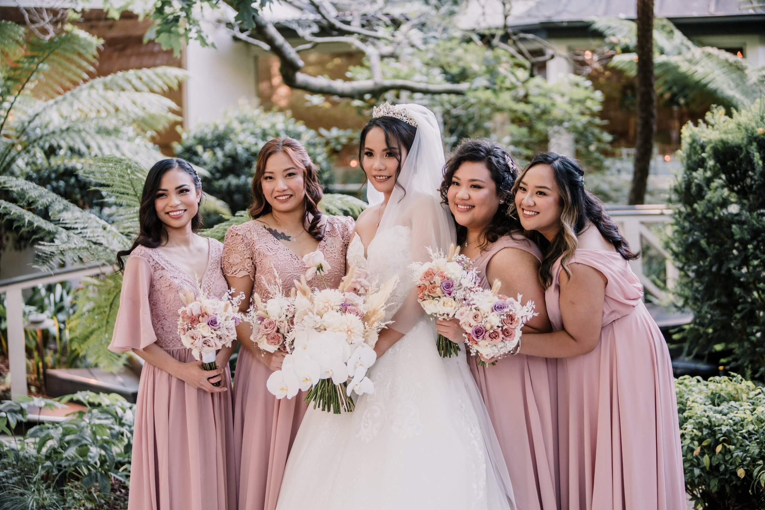 The bride wearing a beautiful ballgown wedding dress and a flowy veil and a tiara is flanked by two bridesmaids on each side while they smile for the camera.