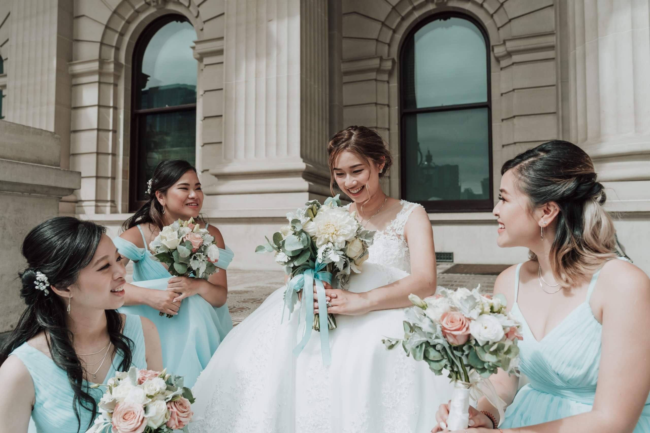 The bride sits on the stairs of a beautiful building with her baby blue-dressed bridesmaids sitting all around her.