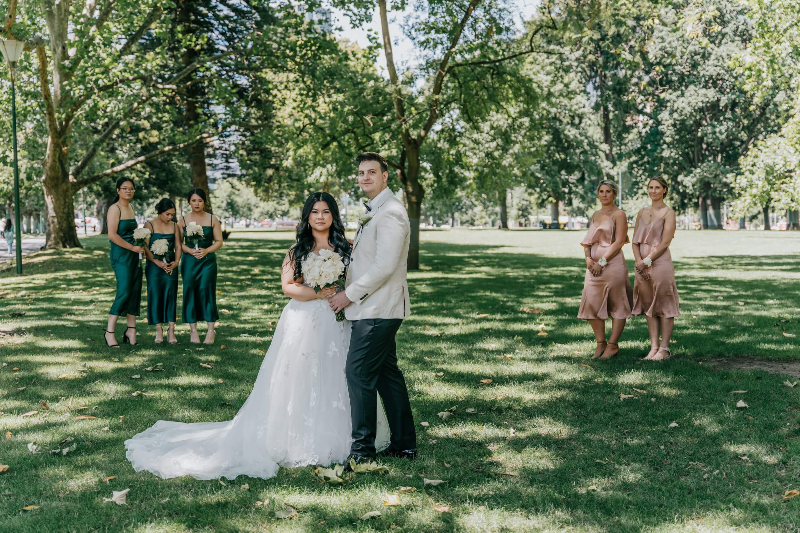 The bride and groom, surrounded by greenery, stands in front of the camera, while 5 bridesmaids wearing emerald green and rose-gold dresses stand in the background