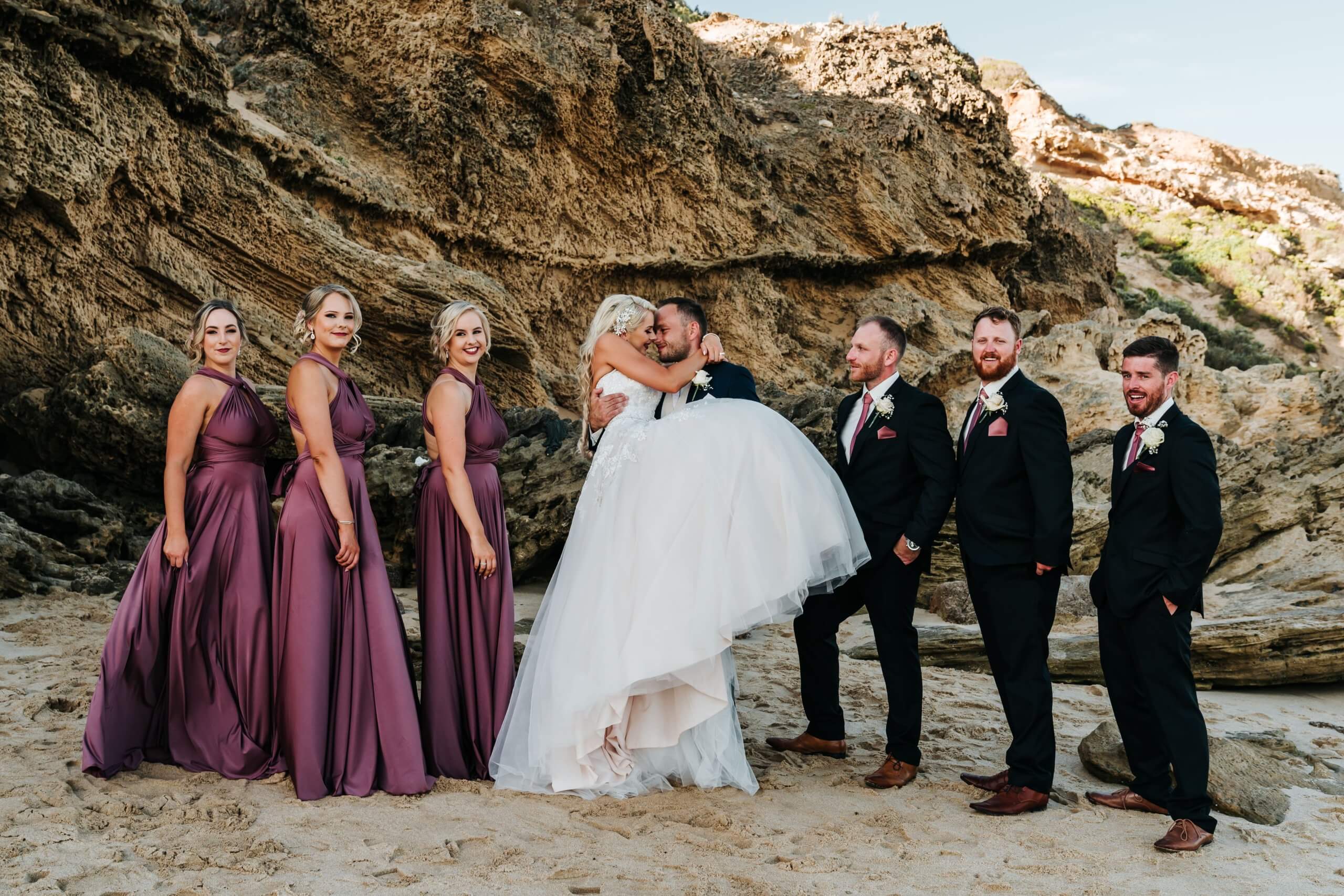 A black and purple-dressed bridal party smiling at the camera, while the groom carries the bride in bridal style.