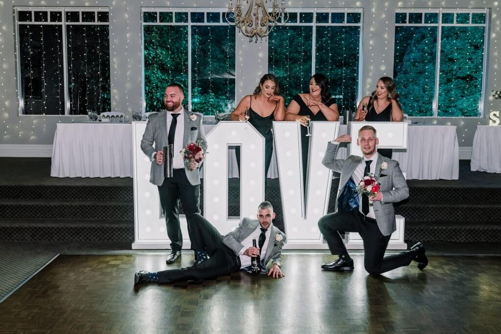 The groom holds a beer mug, posing goofily in front of the Giant L.O.V.E letters with the bridal party
