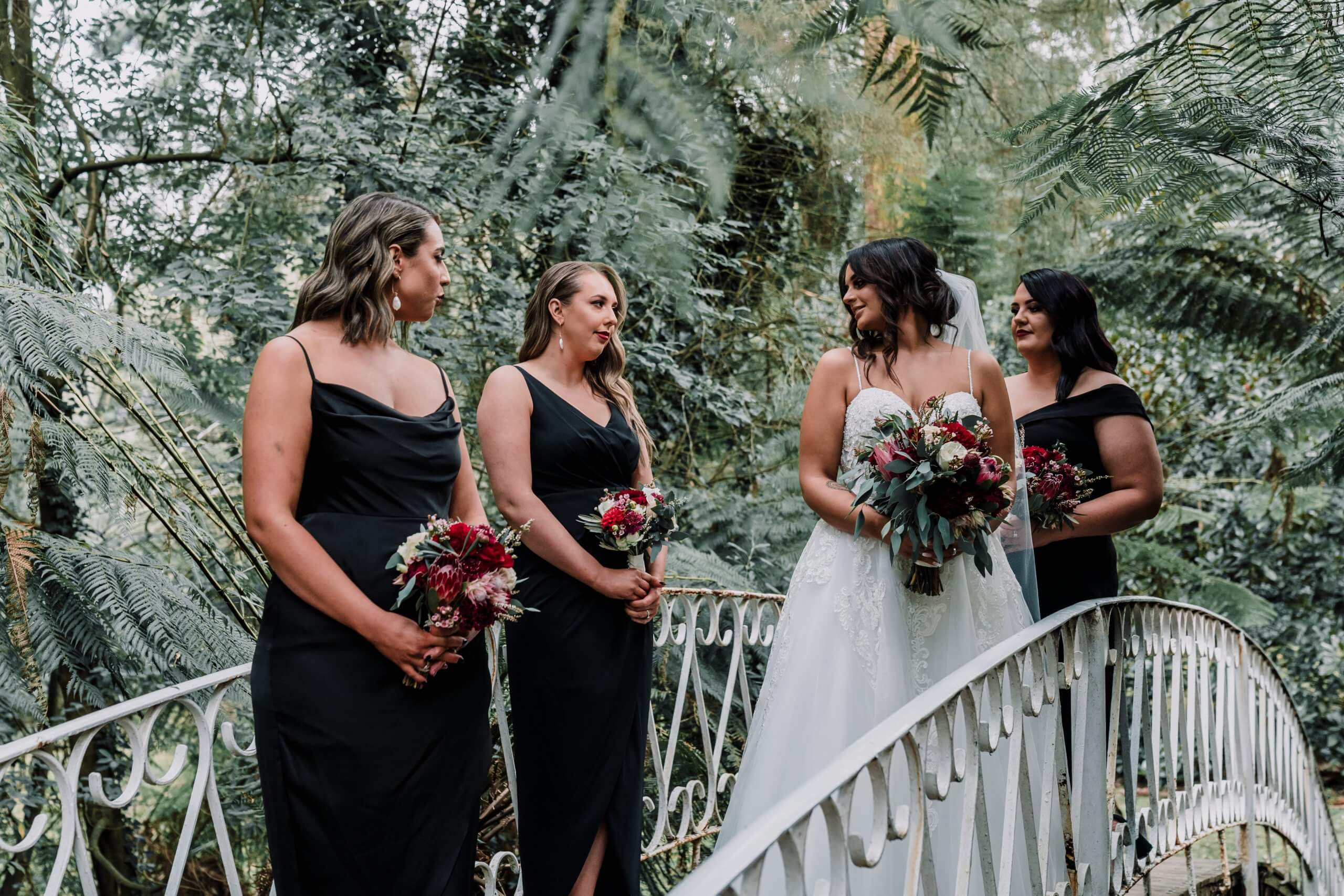 The bride looks stunning in her wedding dress and holding her bouquet of vibrant red flowers. Her three bridesmaids - clad in black dresses is photographed standing atop a bridge.