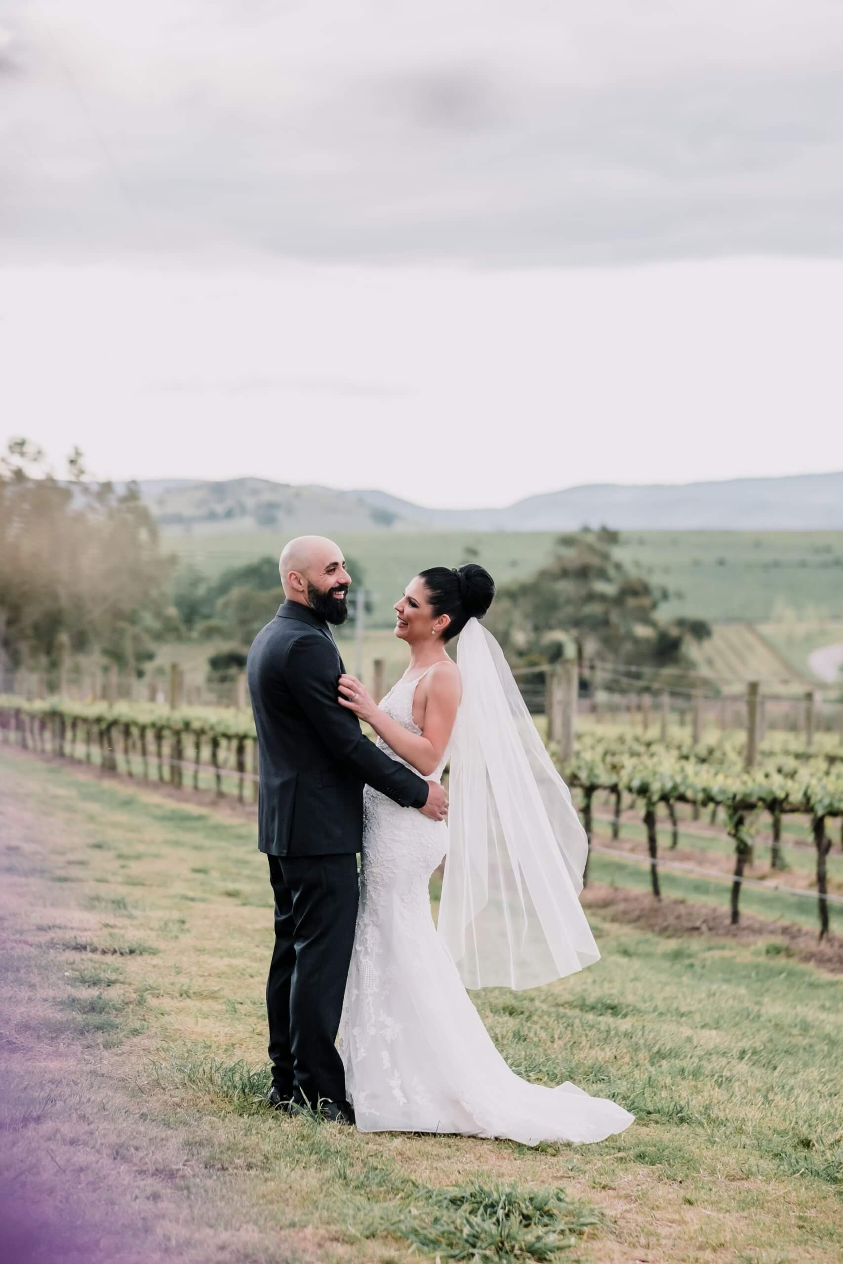 Bride and groom stands facing each other, smiling warmly in the middle of a vineyard.