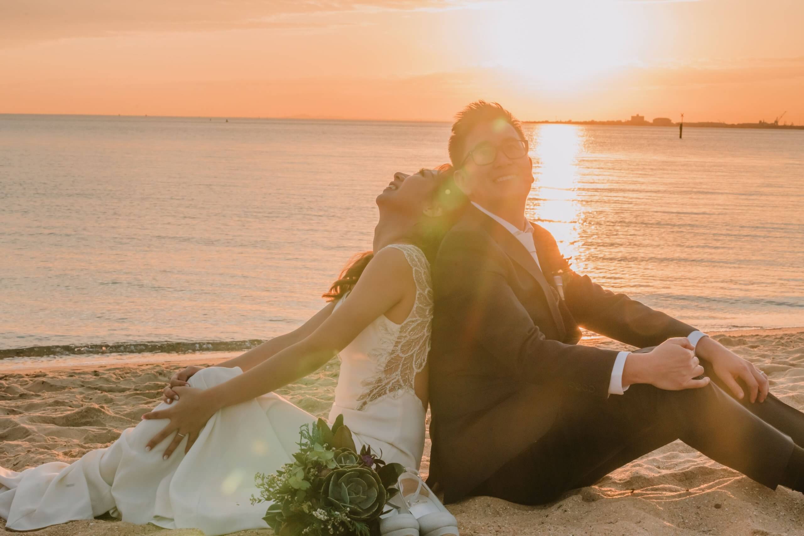 Bath in the golden light of the setting sun, the bride and groom candidly laughs as they sit on the sand and lean their backs on each other.