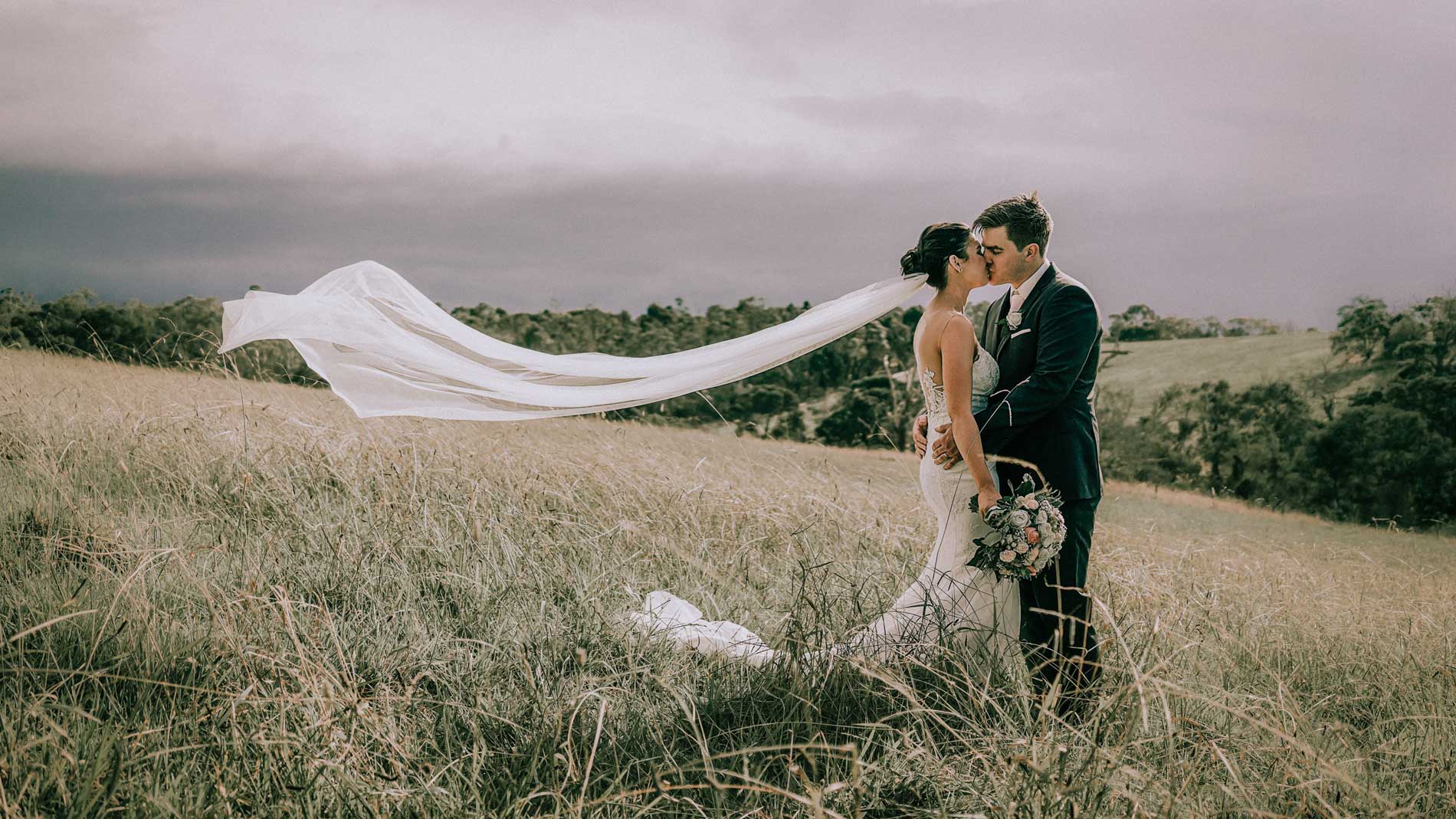 Cinematic photo of the bride and groom as they kiss in the middle of a grass field, with the wind blowing the bride's veil.