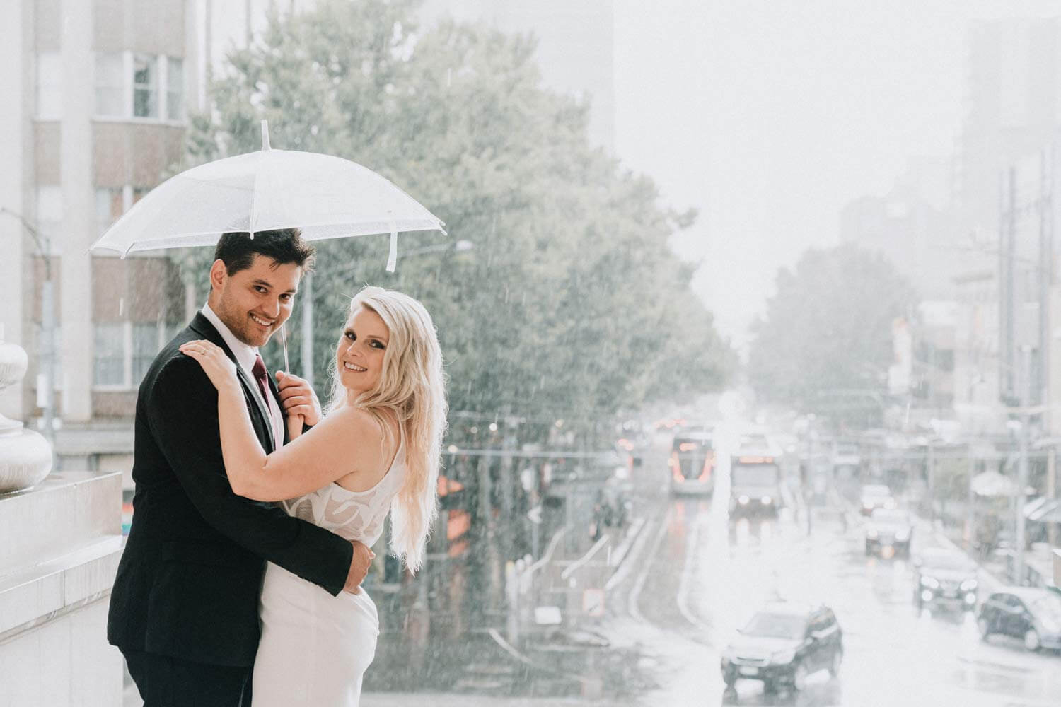 The bride and groom have beautiful smiles on both their faces as they embrace each other, sharing an umbrella under the pouring rain.