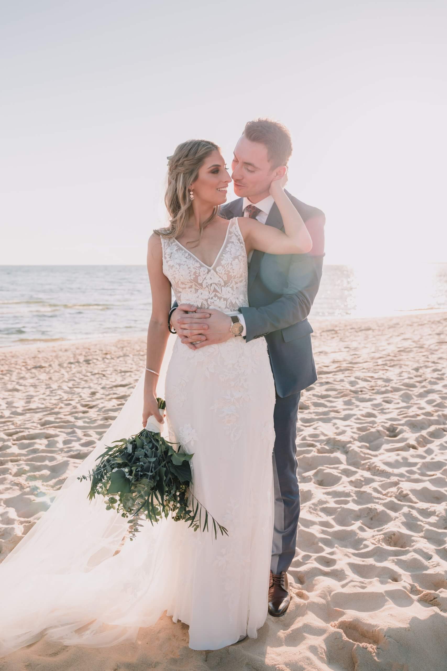 White sand, the setting sun, and the calm waters create a beautiful backdrop for the groom who embraces his bride with both ands on her waist.