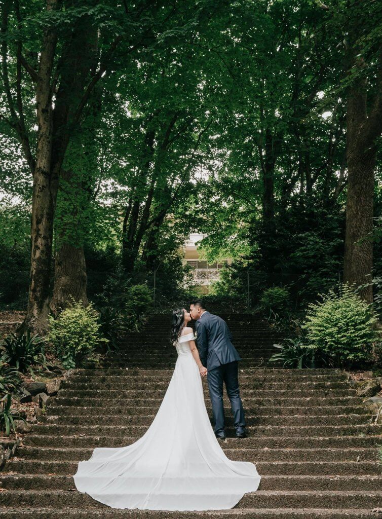 A sweet and beautiful of the bride and groom kissing as they are standing atop a forest-lined staircase. The bride's gown creating a beautiful and magical trail.