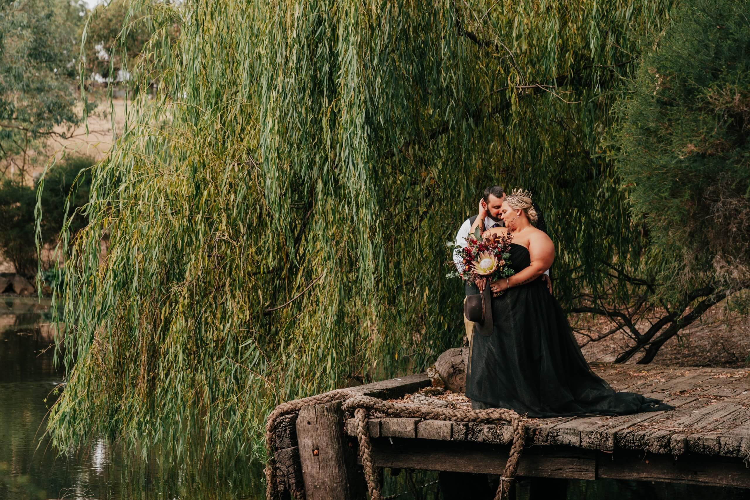 The bride, wearing a black, unconventional wedding gown and a beautiful tiara is being embraced lovingly in back hug by her husband as they are standing in the docks.