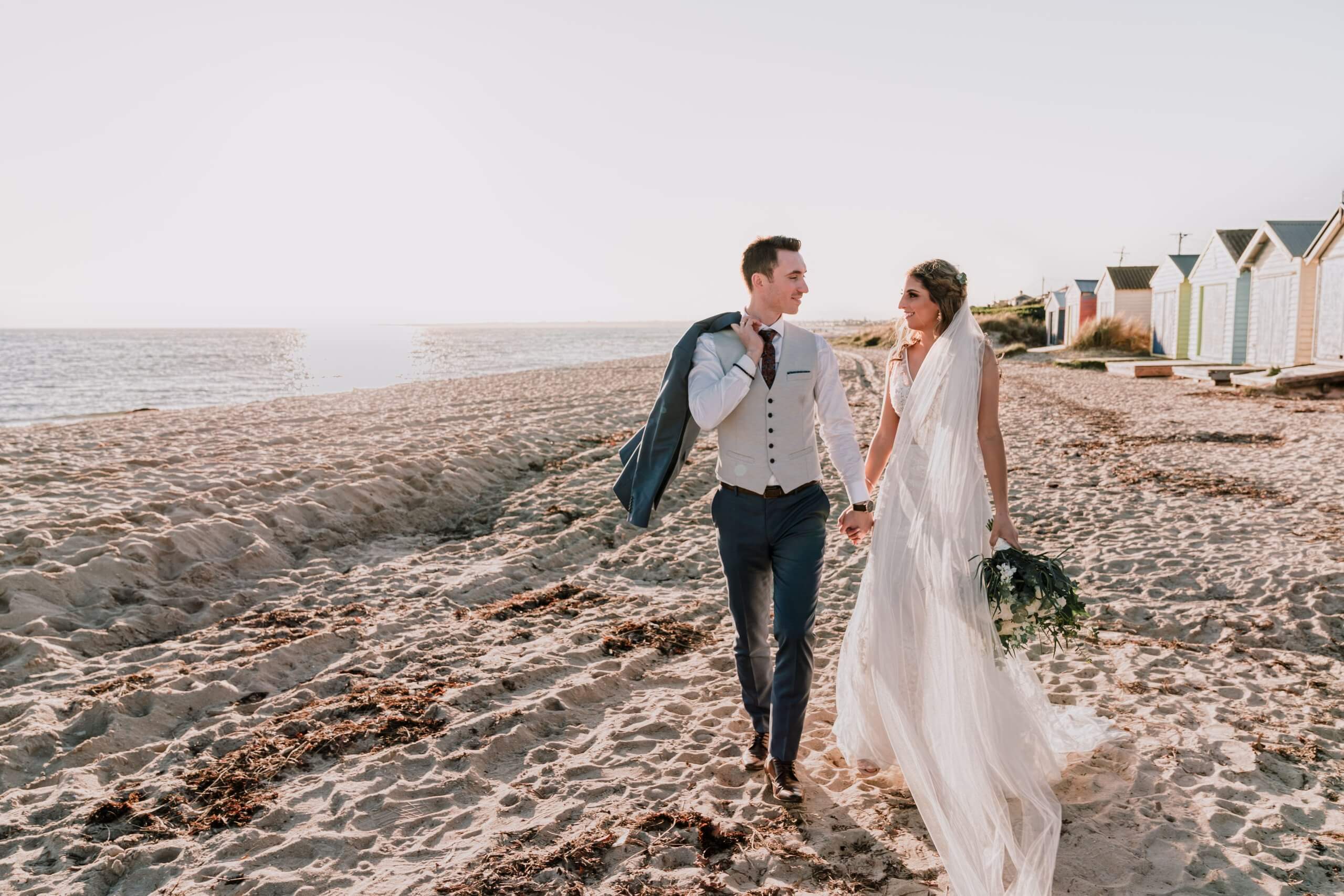 Bride and groom walking the shoreline, with the setting sun casting them in a golden glow.