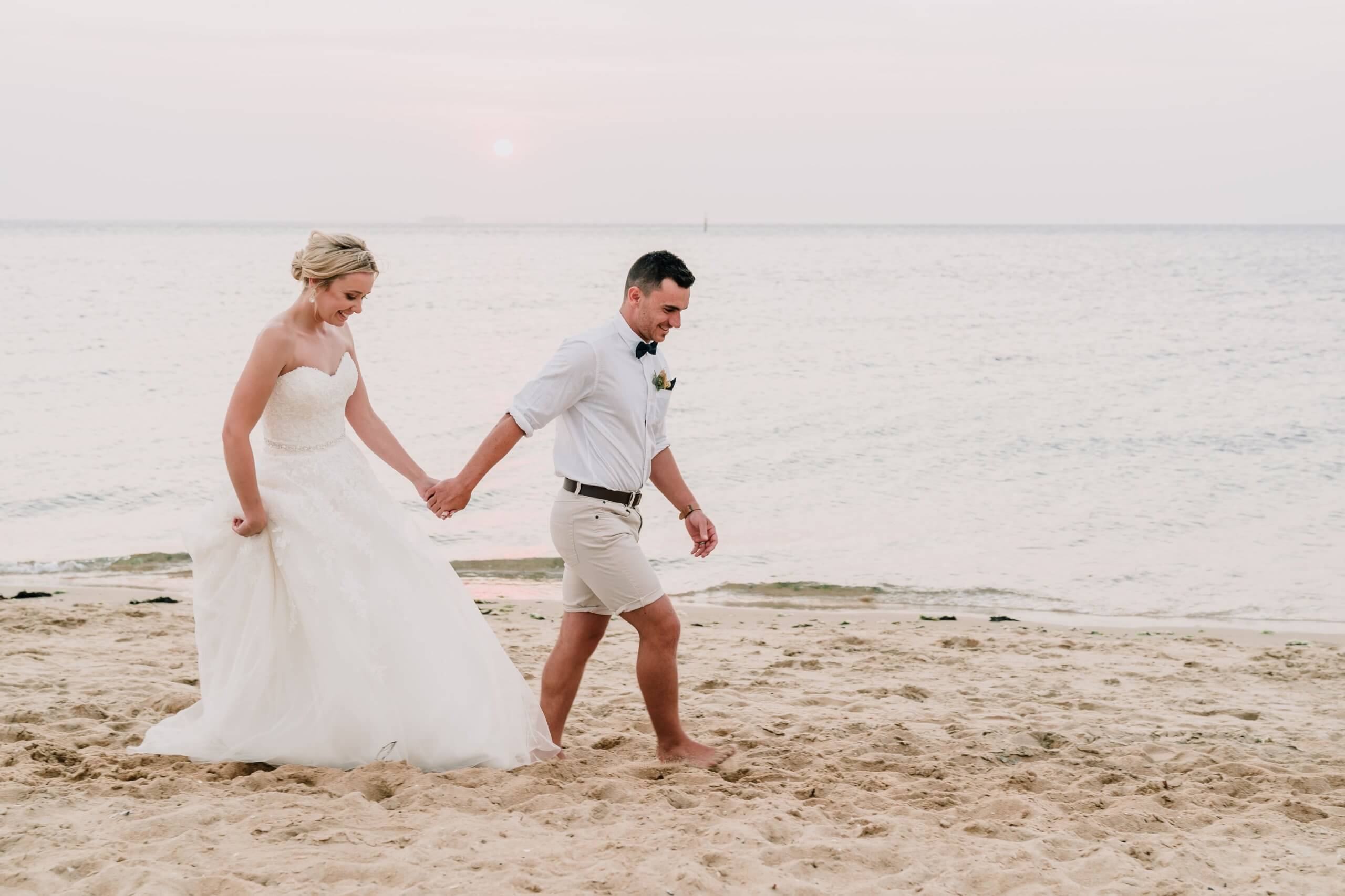 Bride and groom walking hand in hand in the shoreline with the calm sea and the pinkish horizon as the background.