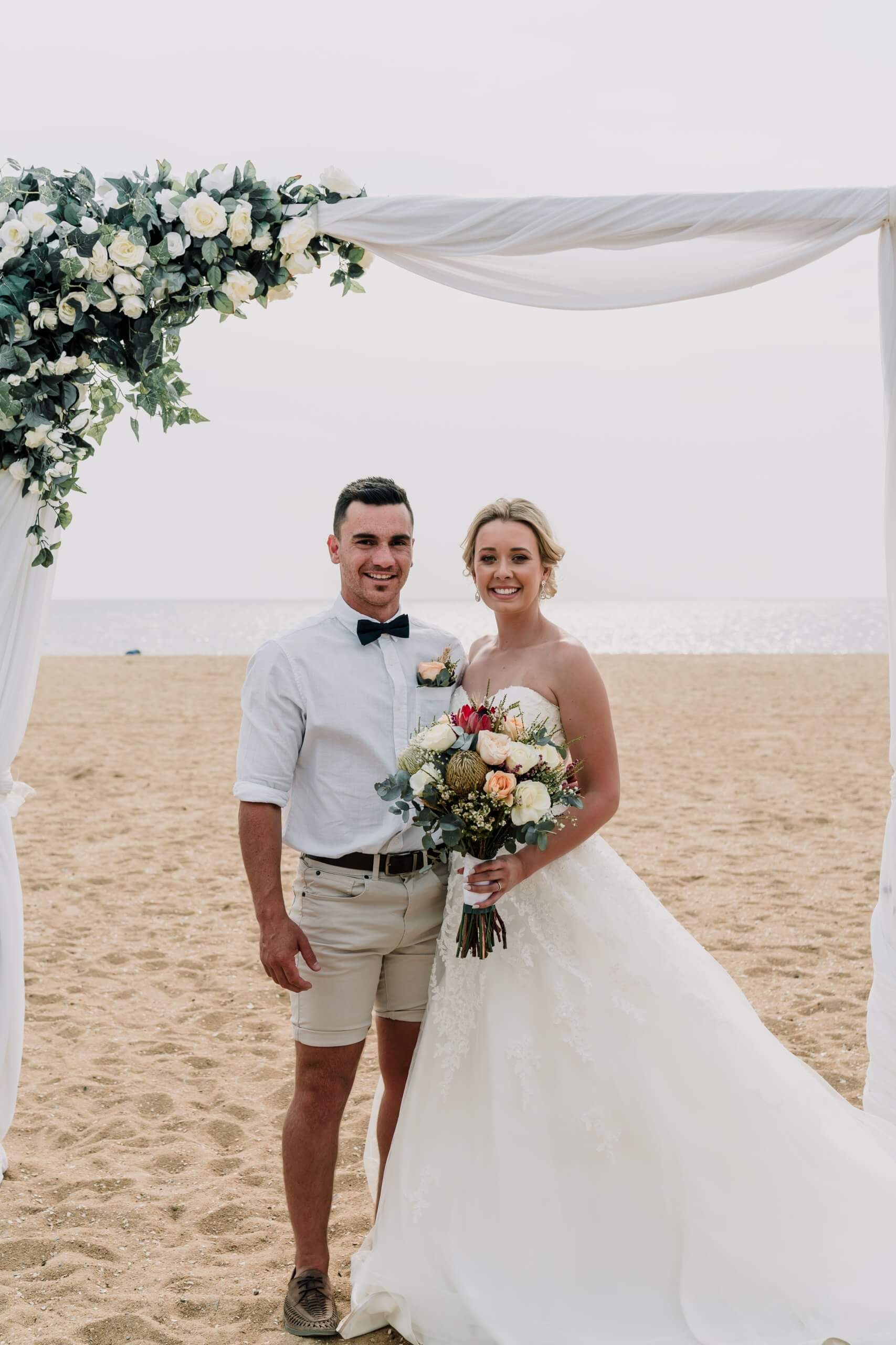 Portrait photo of the bride and groom under the wedding arch at their beach wedding.