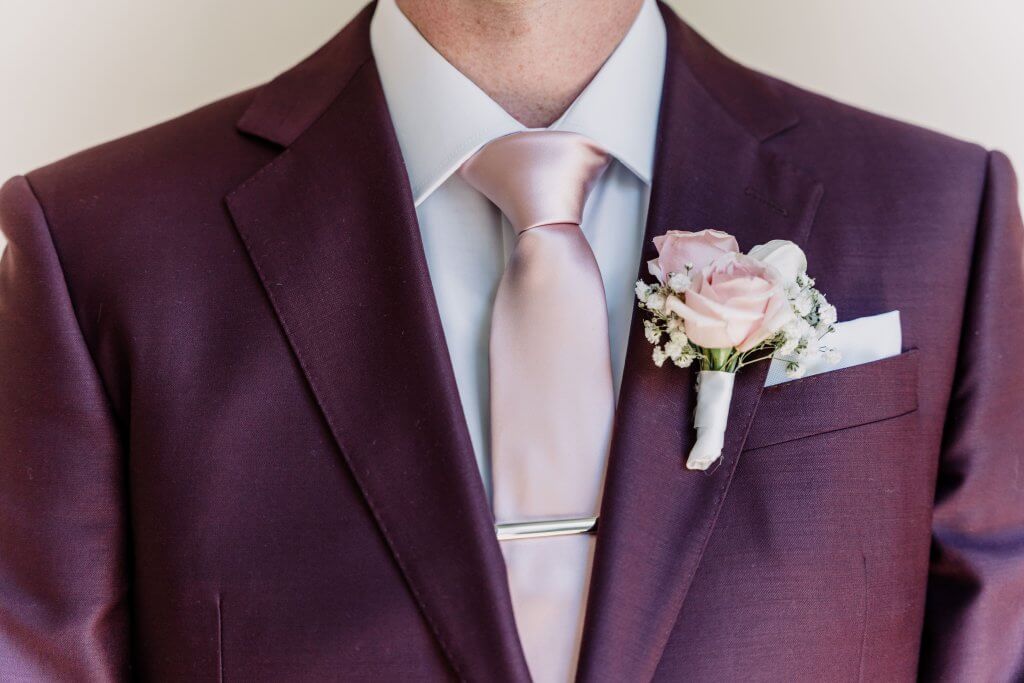 Up close shot of the groom showing his his maroon wedding suit, pink silk tie and pink roses and white babies breath boutonniere.