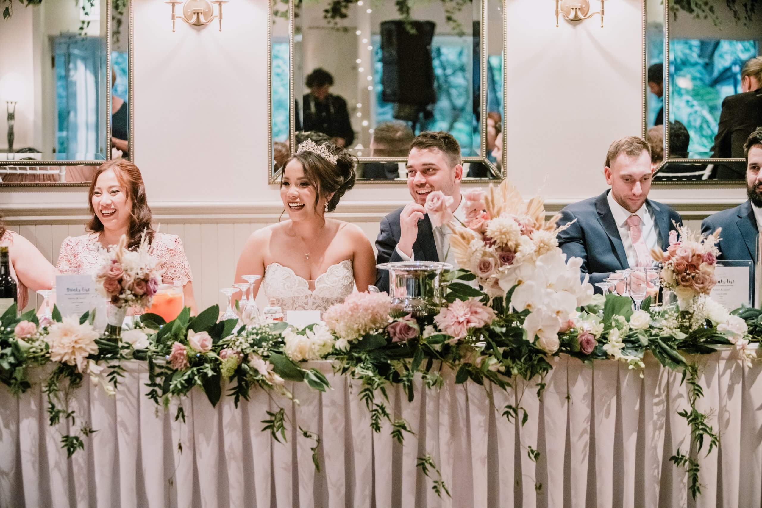 Bridal party sitting on the long table adorned by beautiful flowers and sharing a good laugh.
