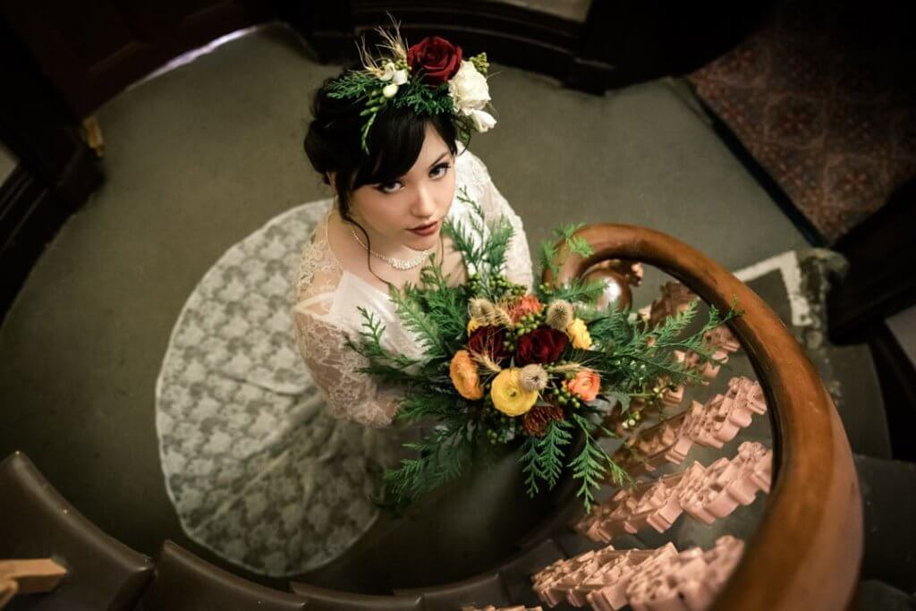 Cinematic photo of the bride looking up at the camera in a staircase while holding her colorful bouquet close to her chest.