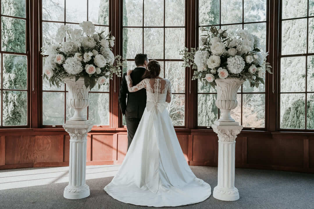 Beautiful photo with the back view of the bride and groom facing large crystal windows and flanked by floral columns on both sides.