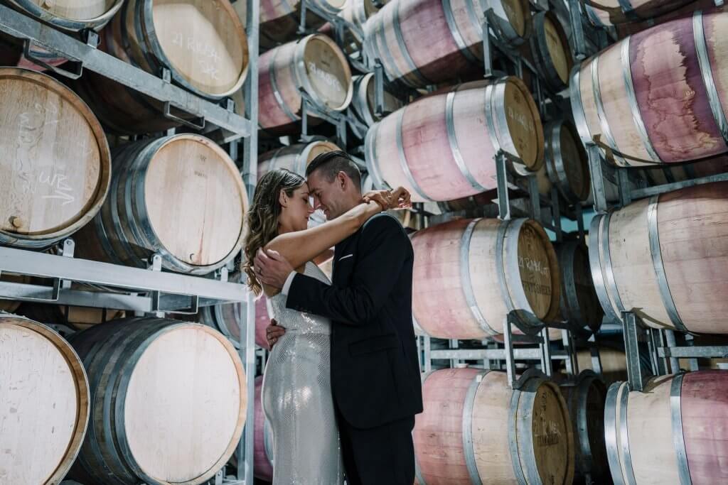 Bride and groom embracing each other lovingly while they are standing in the middle of a warehouse filled with barrels of wine.