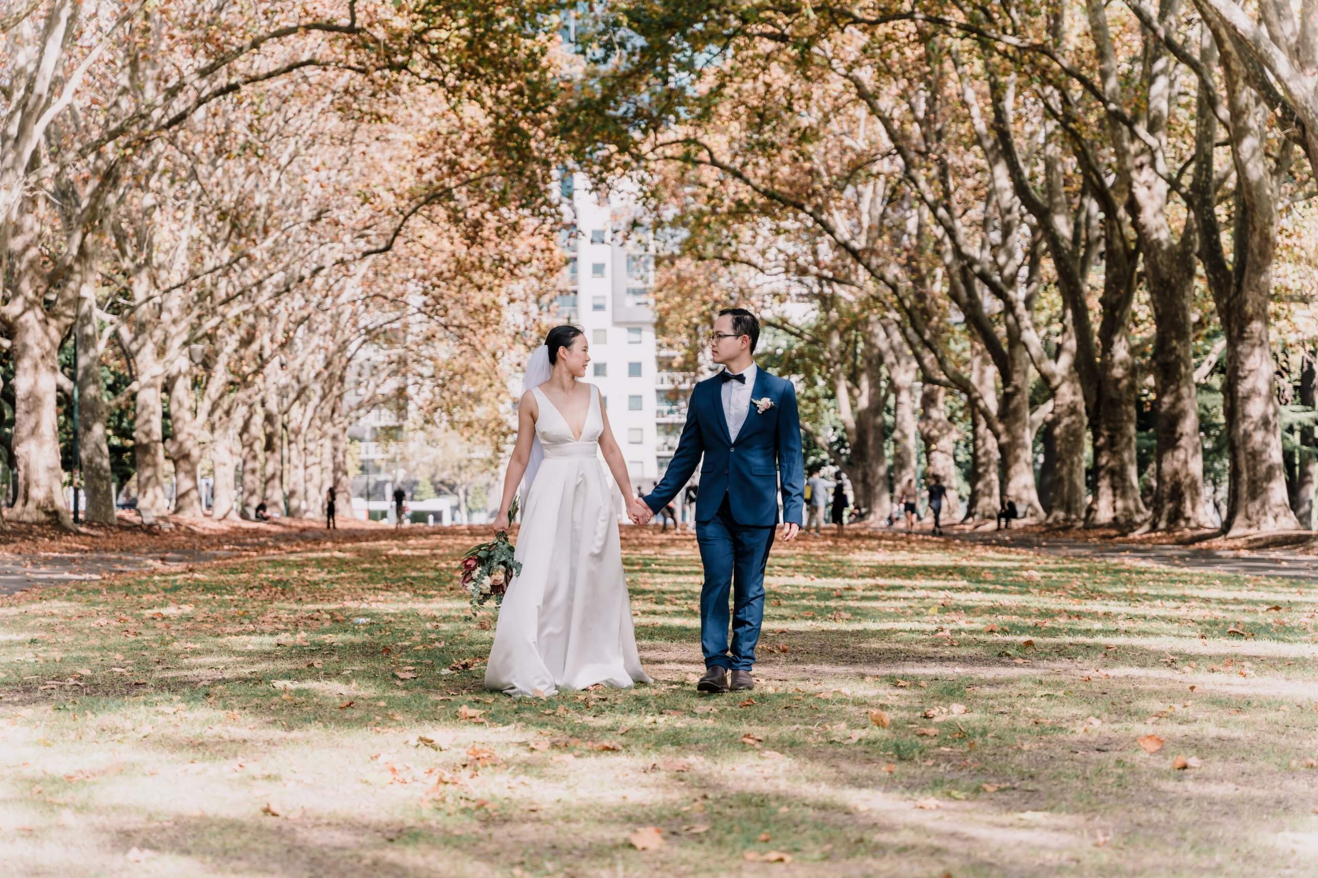 Big, tall trees line the park where the bride and groom stands in the middle holding hands.