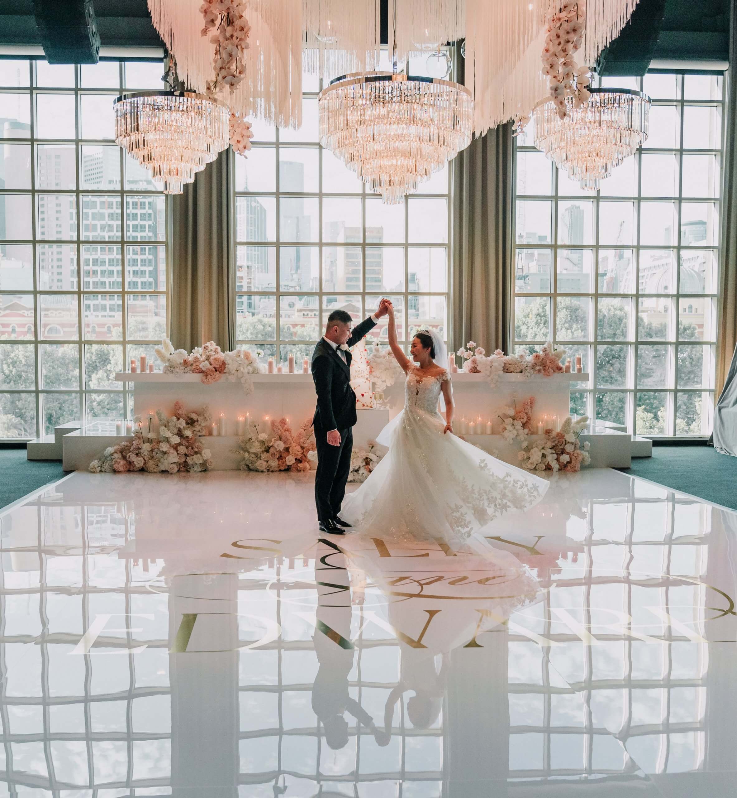 Stunning photo of the bride and groom during their first dance. The groom twirls the bride as they dance surrounded by pretty pink flowers and under a stunning crystal chandelier.