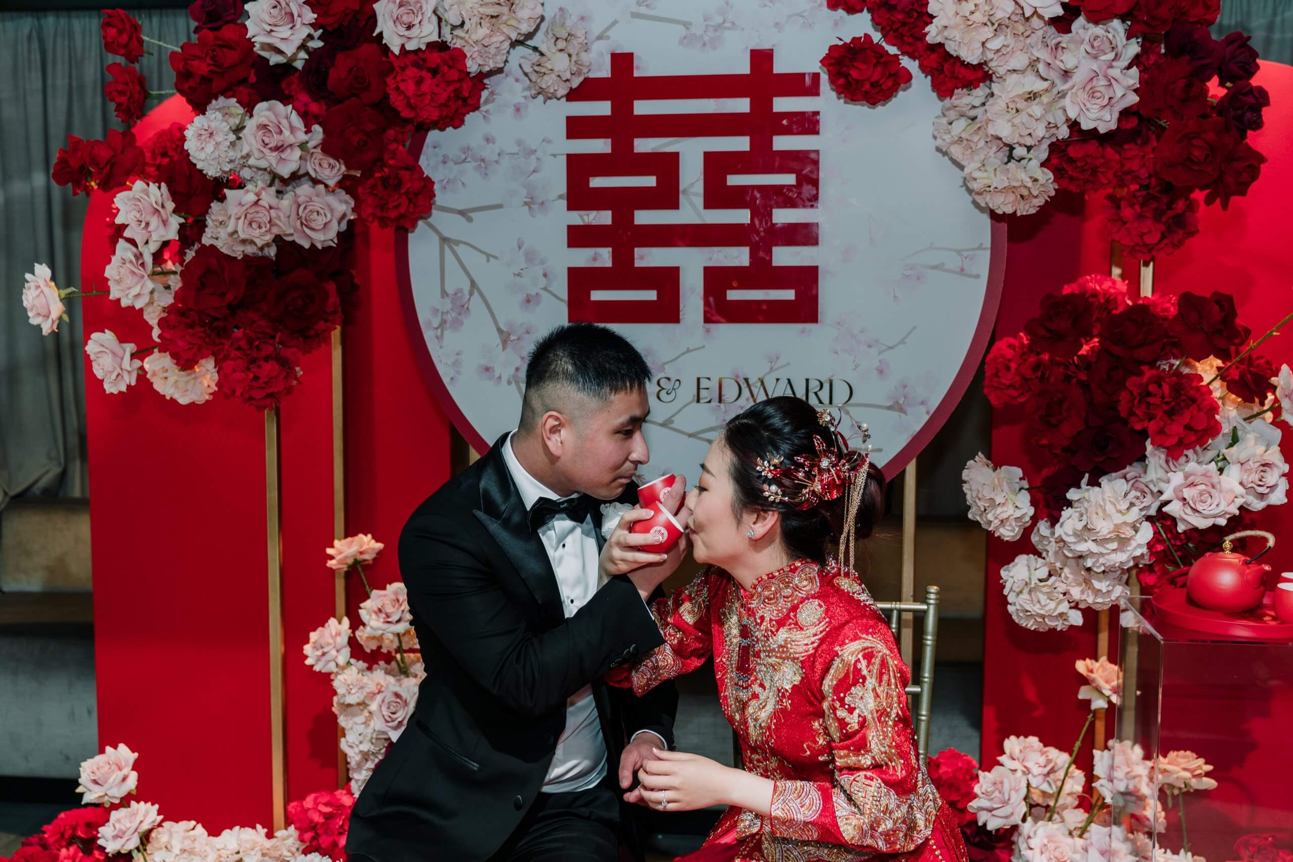 Customise Your Wedding - During their tea ceremony, surrounded by pretty red and pink flowers, the bride (wearing a red traditional Chinese dress) and the groom (black and white tux) drink tea in a toast.