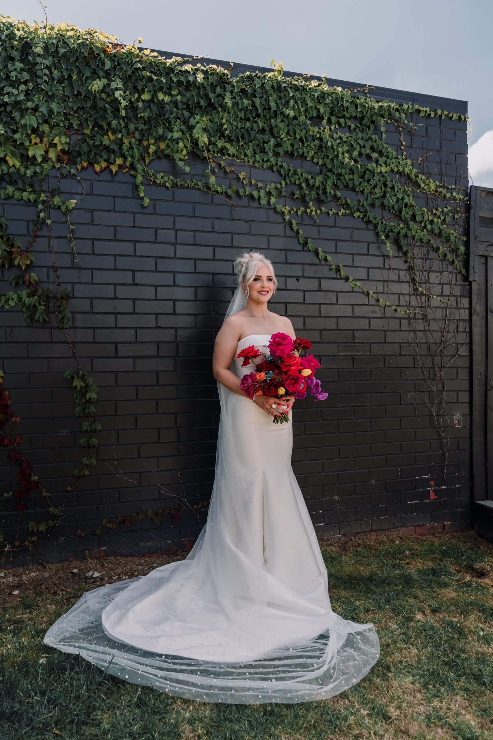 Bride showcases her full wedding gown while holding her beautiful bouquet of bright red and pink roses.