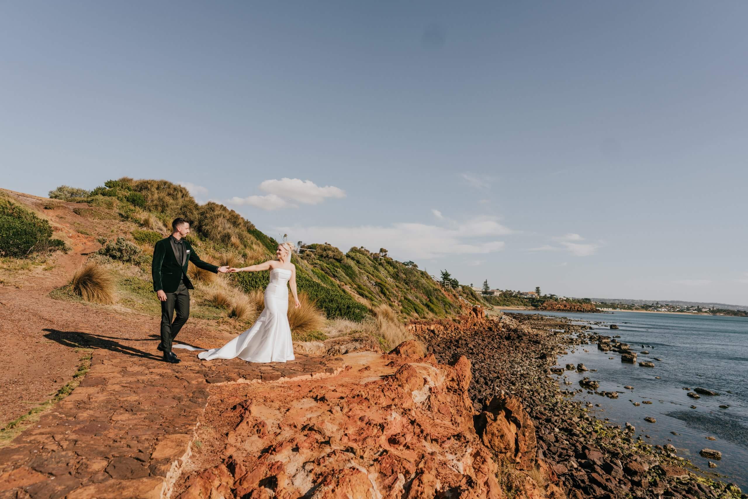 Bride and groom holding hands at the rocky Moonah Links shore