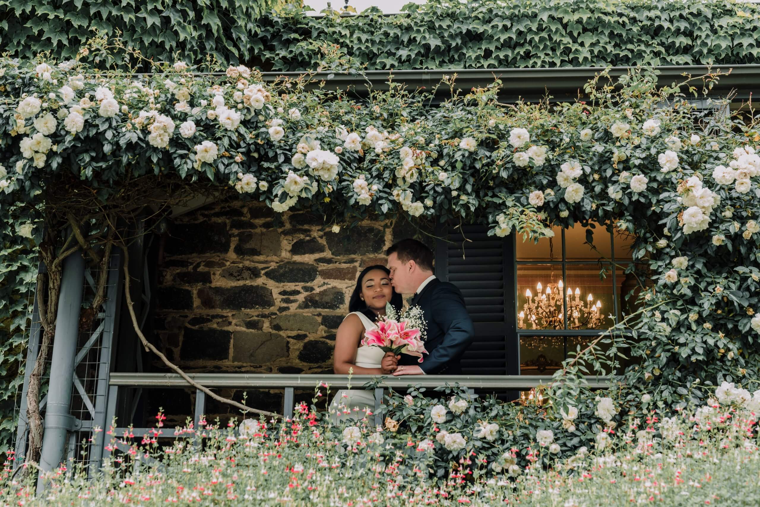 The groom kissing the bride's cheek as they stand behind a heart-shaped hanging bush at Overnewton Castle